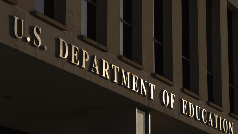 Light reflects of a letter sign reading U.S. Department of Education affixed to the side of a stone masonry building beneath a row of tall windows.