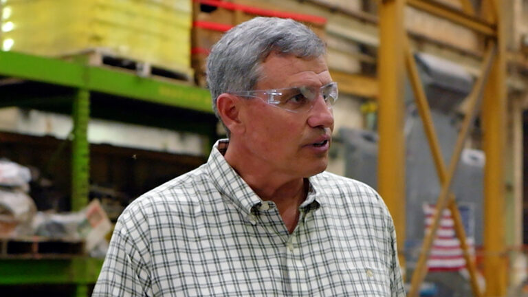 Bill Berrien speaks while wearing safety glasses and standing in a warehouse, with out-of-focus metal shelves filled with pallets of materials in the background.