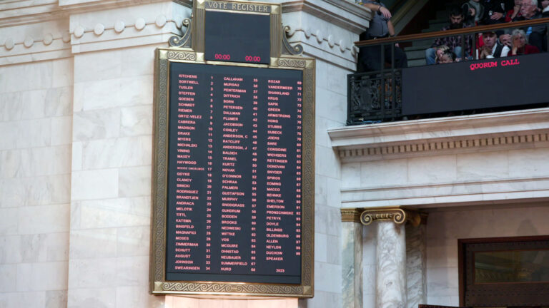 A digital vote register mounted on a marble-block masonry wall shows names and district numbers, with another digital sign displaying the words Quorum Call mounted on a metal grille fence with a wood railing in front of a terraced gallery with seated people, in a room with marble masonry and pillars.