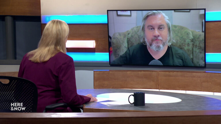 Frederica Freyberg sits at a desk on the Here & Now set and faces a video monitor showing an image of Adam Kunz.