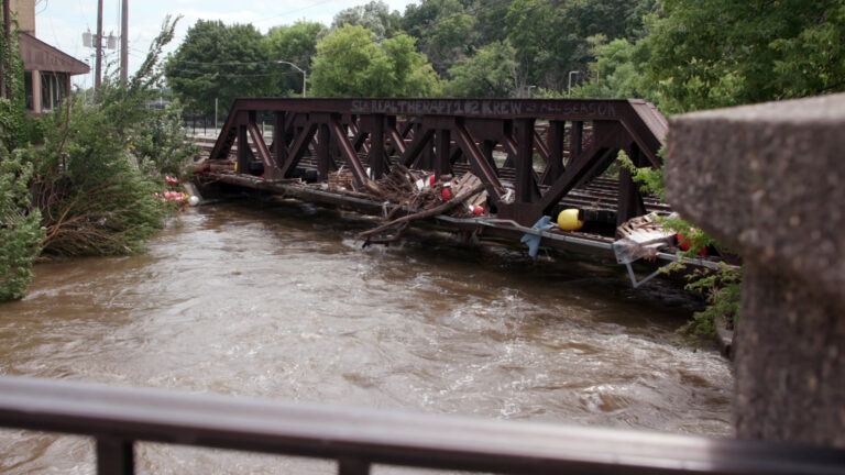 Silt-laden water flows close to the bottom of a railroad bridge with pieces of trees, wood pallets and other debris piled on its surface, with bend trees on one shore, a line of trees, utility lines and a building in the background, and an out-of-focus metal railing and concrete pillar in the foreground.