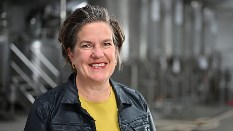 Missy Hughes poses for a portrait while standing in front of an out-of-focus row of stainless steel fermentation tanks in a high-ceilinged room.