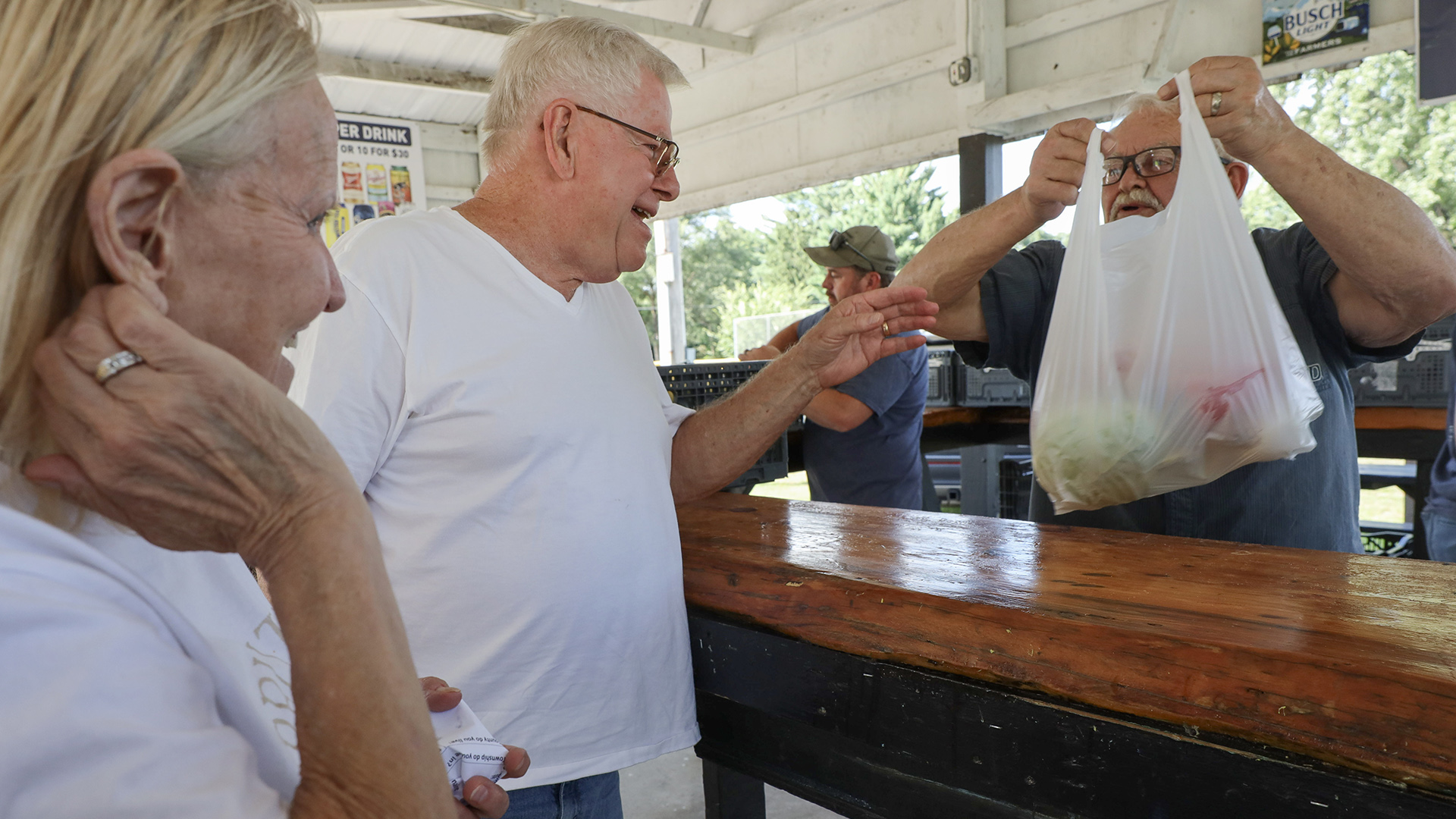 Dave Dearth extends his left hand over a polished wood bar and reaches toward a plastic bag filled with produce held by another man while standing under an open-walled wood structure, with another man reaching into a plastic crate in the background and a woman standing at left in the foreground.
