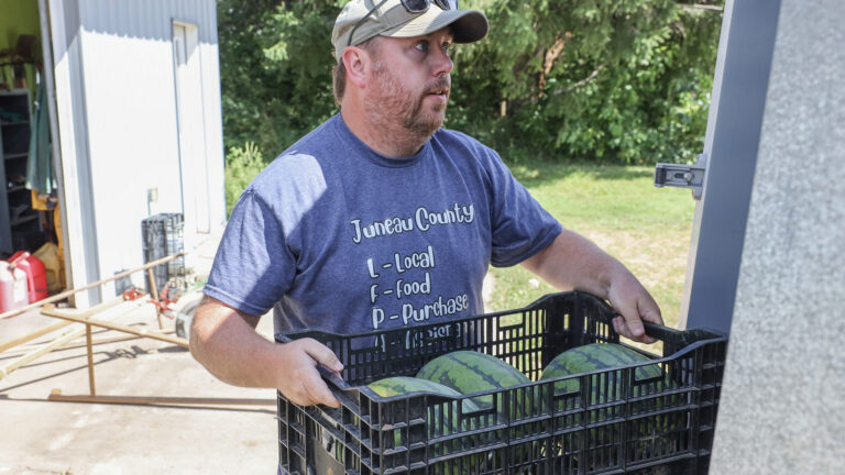 Dustin Ladd holds a plastic crate filled with watermelons while standing next to a door with a lock latch, with a metal building and concrete patio next to a lawn and trees in the background.