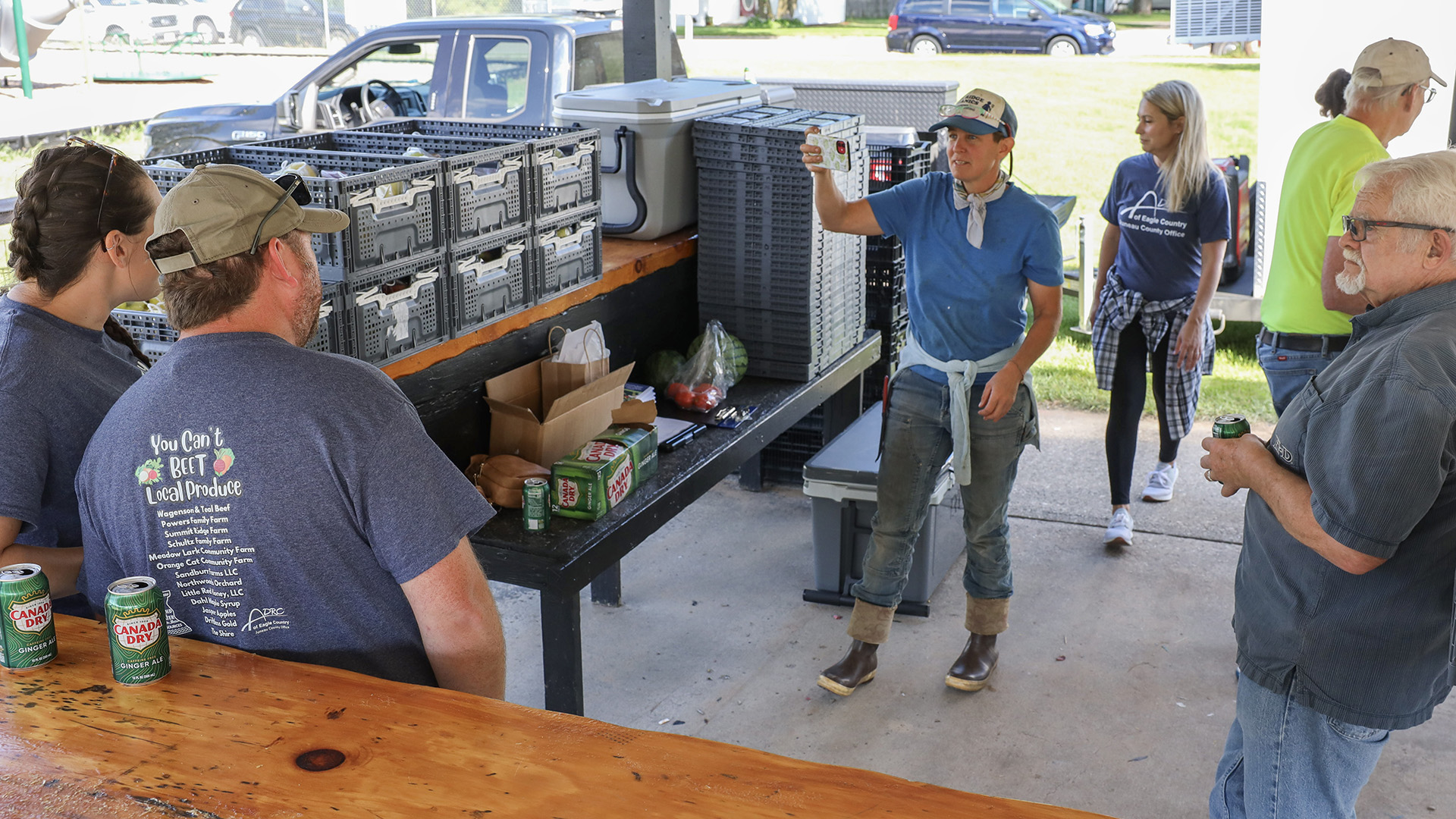 Multiple people stand in between multiple wood benches and bars stacked with plastic crates and soda cans while standing on a concrete surface, with parked vehicles and a lawn in the background.