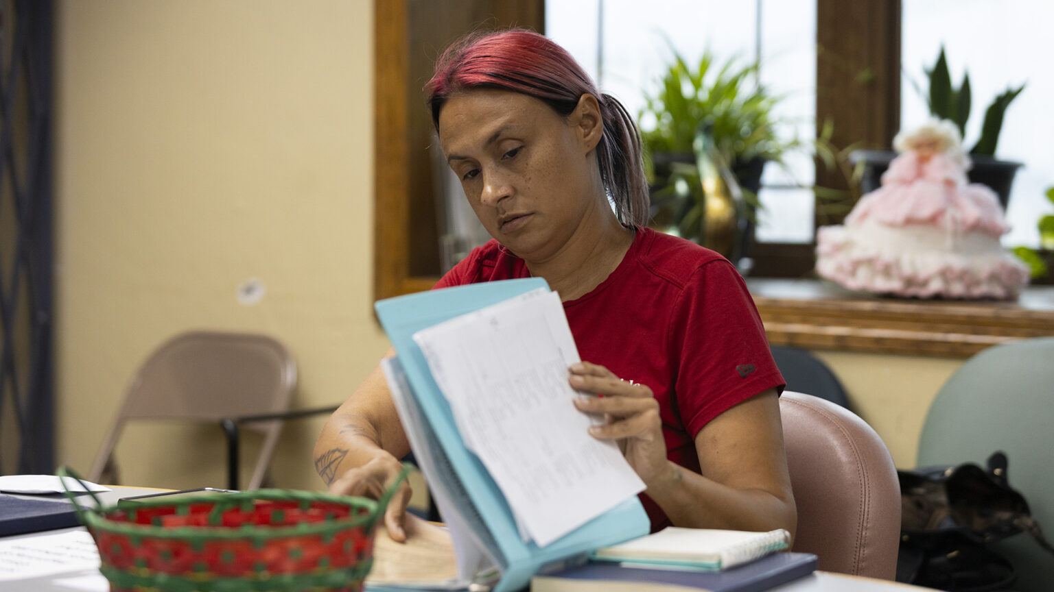 Tracy Germait uses her left hand to hold papers to the front cover of a three-ring binder held open while using her right hand to page through its contents, while seated at a table with books, papers and a basket on its surface, in a room a row of chairs along a wall under a windowsill with plants and a doll on its surface in front of a closed wood-framed window. Tracy Germait uses her left hand to hold papers to the front cover of a three-ring binder held open while using her right hand to page through its contents, while seated at a table with books, papers and a basket on its surface, in a room a row of chairs along a wall under a windowsill with plants and a doll on its surface in front of a closed wood-framed window.