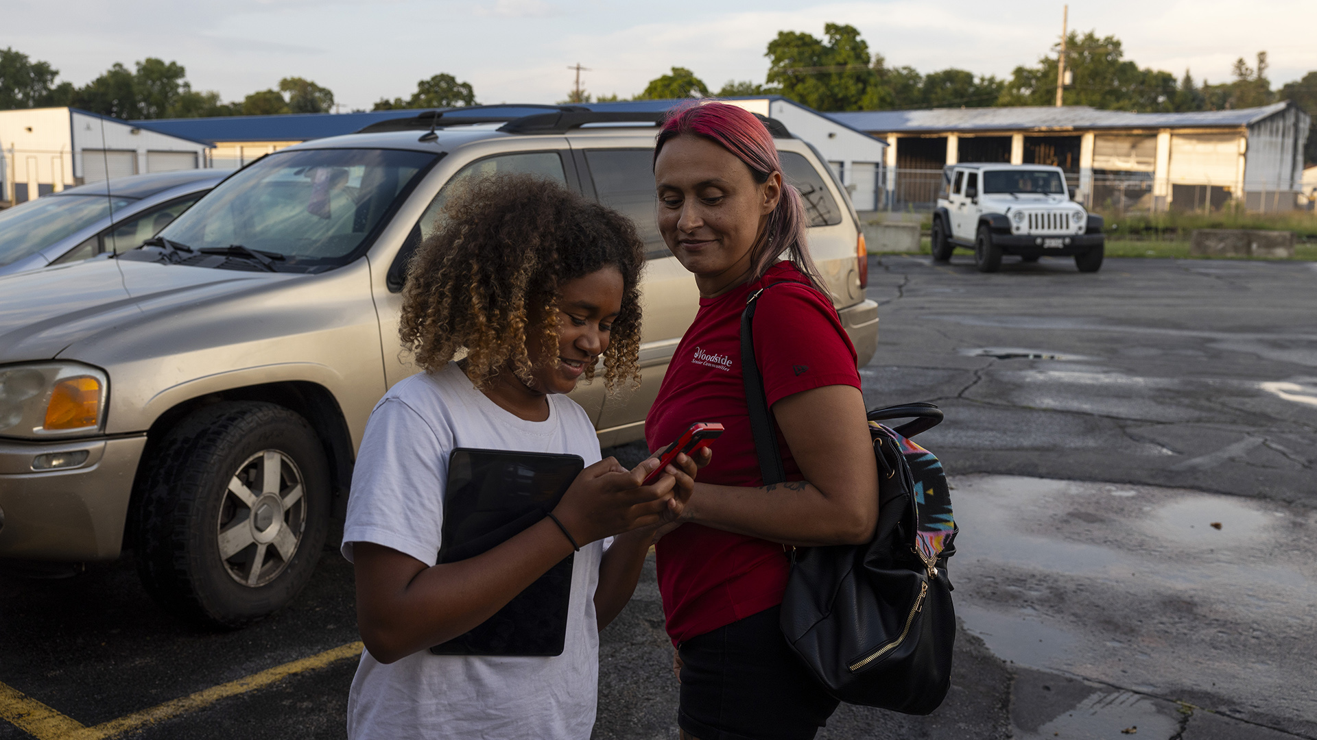 Tracy Germait looks over the shoulder of Isis, who is viewing a phone in her left hand and holding a table to her side with her right forearm, while standing outside in a parking lot in front of multiple vehicles, with storage locker buildings, utility poles and trees in the background.