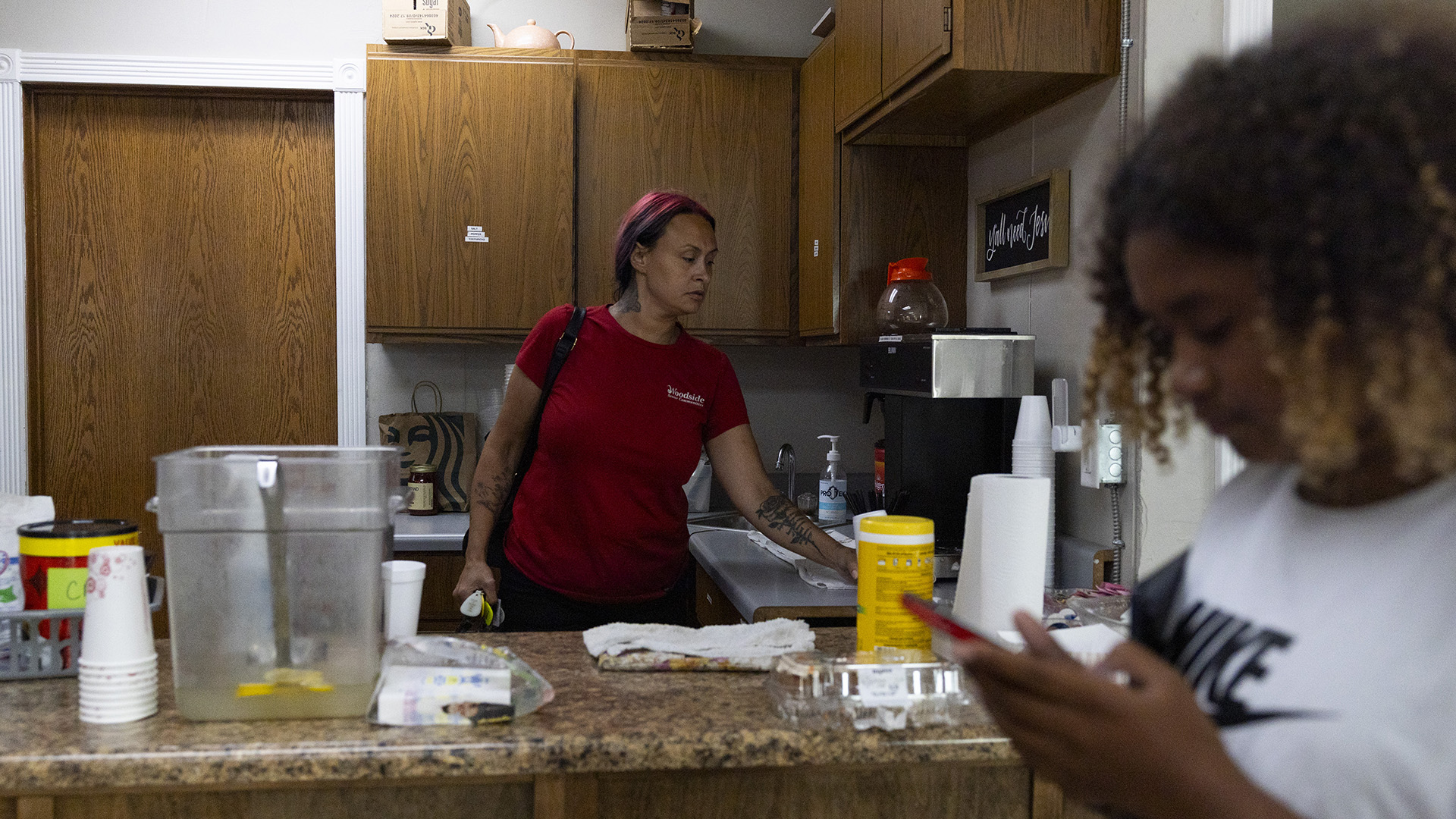 Tracy Germait wipes a counter with a towel while standing in a kitchen behind another counter with paper cuts, towels, plastic containers and other items on its surface and in front of a row of cabinet and a closed wood door, with Isis holding a phone in her hands while standing out-of-focus in the foreground.
