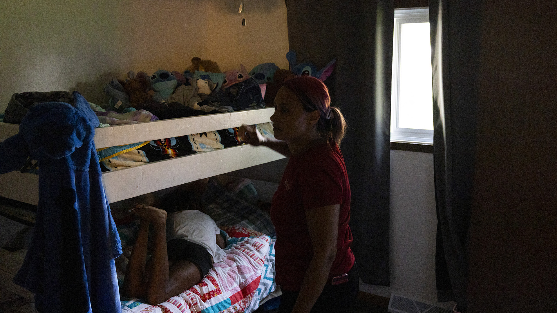Tracy Germait stands next to a bunk bed, with multiple stuffed animals on its top bunk and Isis laying face down on the bottom bunk in a room with partially closed curtains in front of a window.