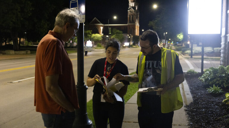 Jamie Wall faces Lucia Sanchez and Cody Oberhuber as they stand outside at night on a sidewalk next to an intersection of two streets, with a mulched garden bed and illuminated sign behind them and more trees, street lights and a brick church with a steeple in the background.
