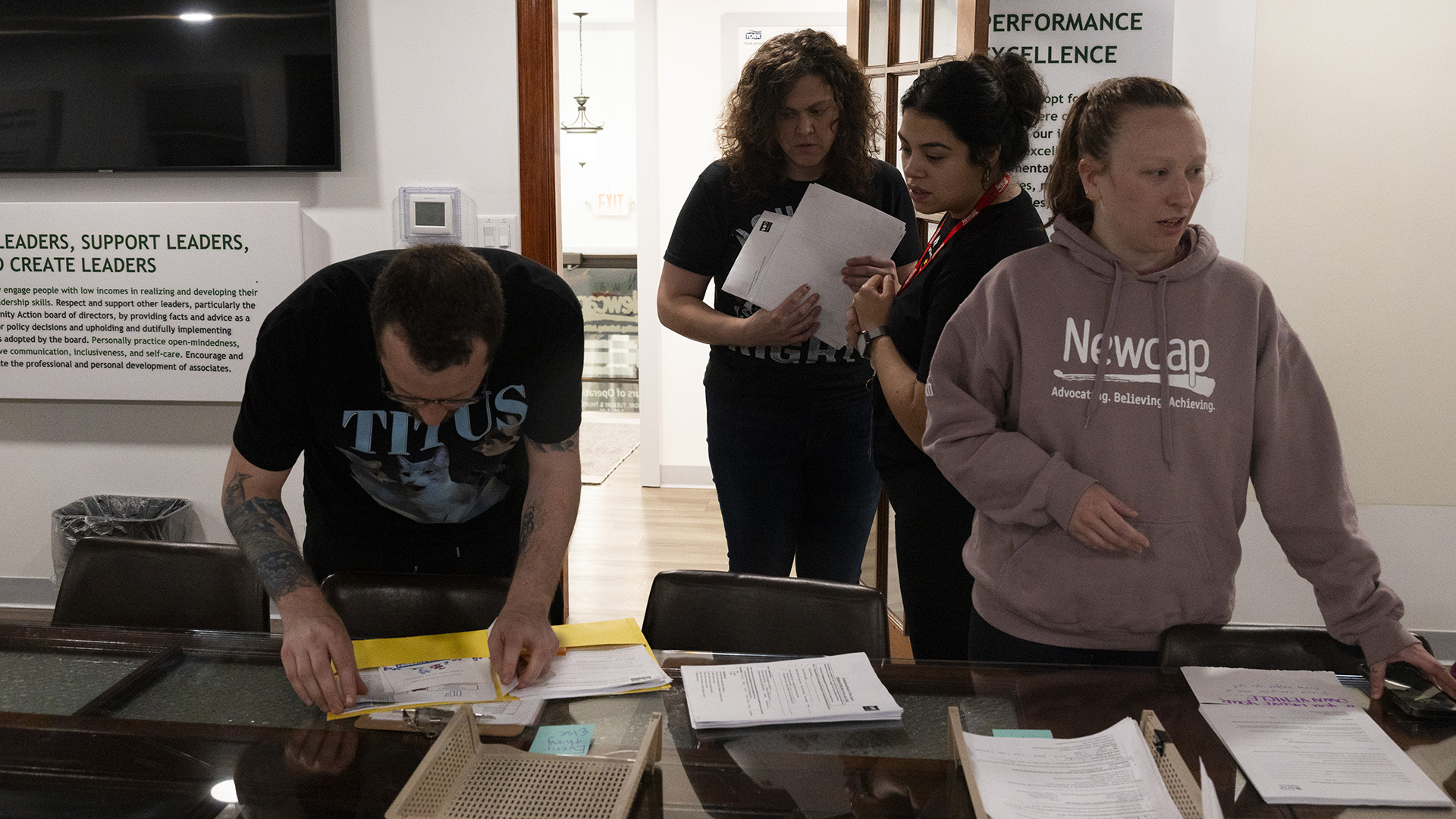 Cody Oberhuber and Alexandra Richmond stand behind multiple chairs and a table with multiple stacks of paper on its surface, with Meagan Gleason and Lucia Sanchez standing behind them in a room with a wall-mounted television and a door opening to another room.