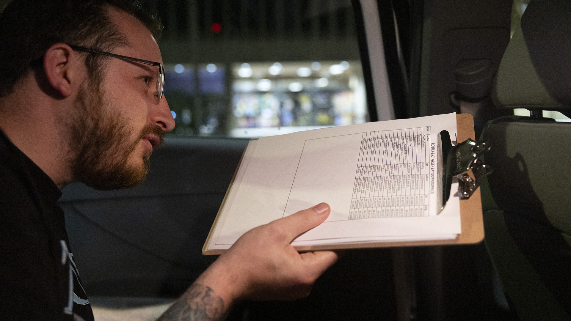 Cody Oberhuber holds a clipboard holding several sheets of paper in his right hand while seated in the rear passenger area of a vehicle, with an out-of-focus storefront visible through a window.