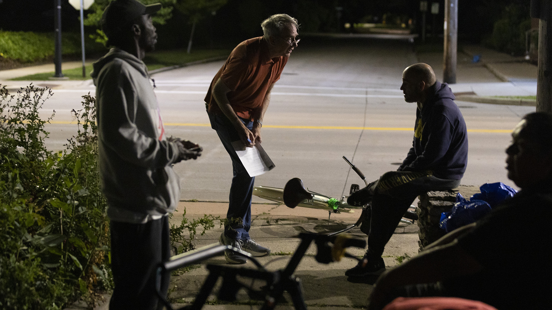 Jamie Wall leans over and writes on a sheet of paper pressed against his right leg facing a man seated next to a bicycle sitting on its side on a sidewalk, with two other men standing and sitting in the foreground next to overgrown foliage, with a street intersection and sidewalks in the background.