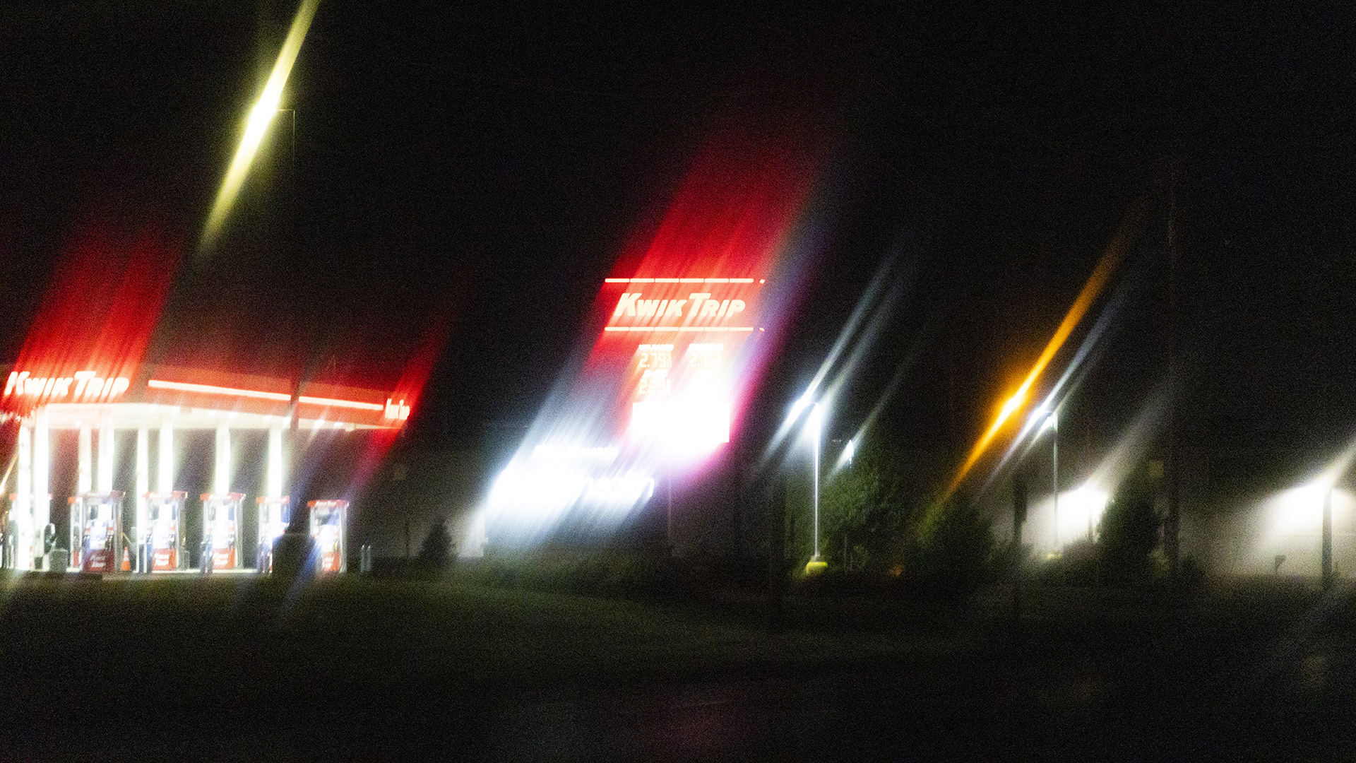 Light from Kwik Trip signs on a pole and above a series of gas pumps along with multiple street and building lights streak across a night sky in a blurry photo.