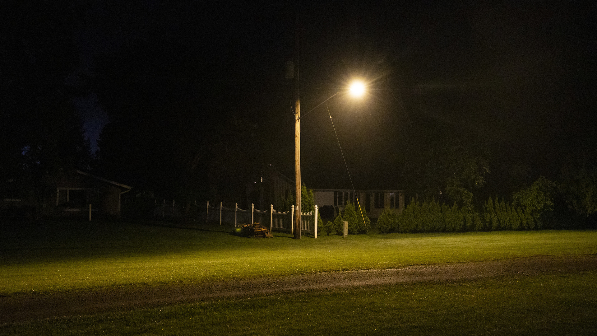 A streetlight next to a picket fence between two houses and next to a line of short coniferous trees illuminates a lawn with a dirt track at night.