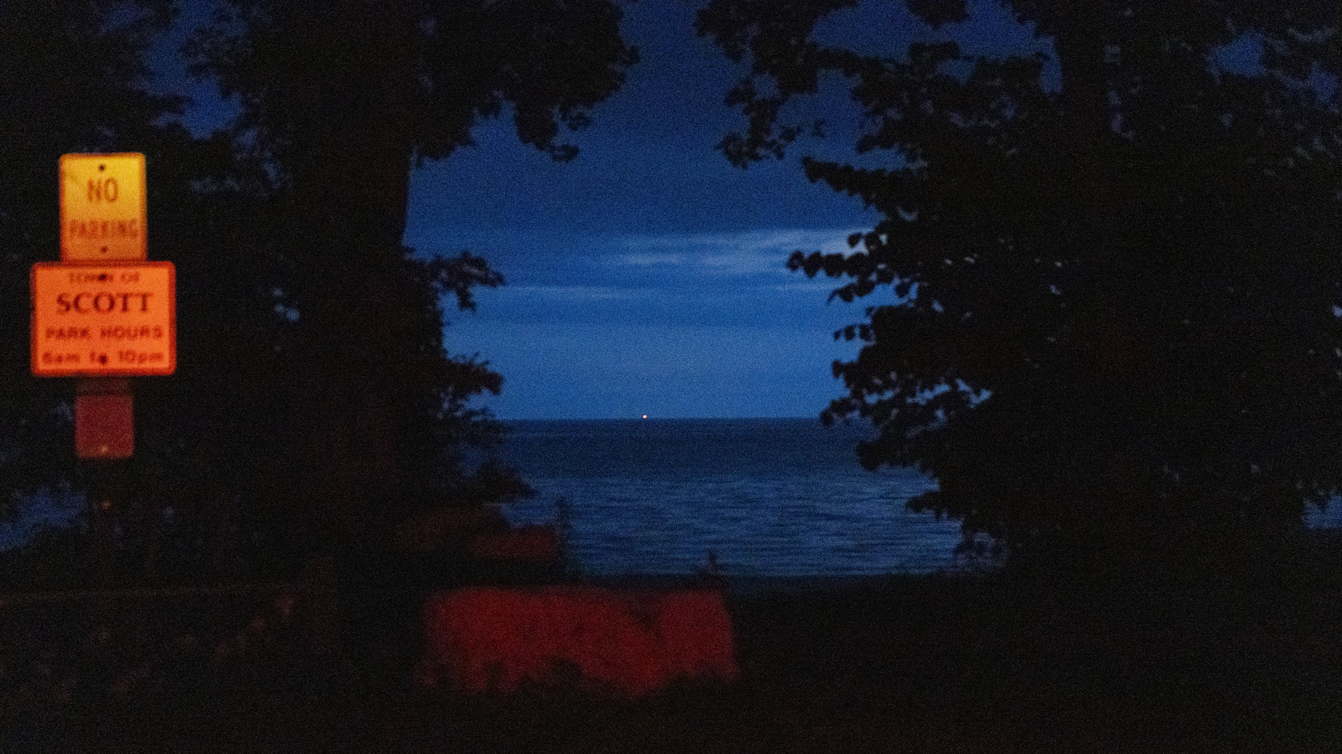 Vehicle headlines illuminate a "No Parking" and park hours sight standing in front of trees along a beach, with a point of light visible on the horizon of a body of water under a cloudy sky.