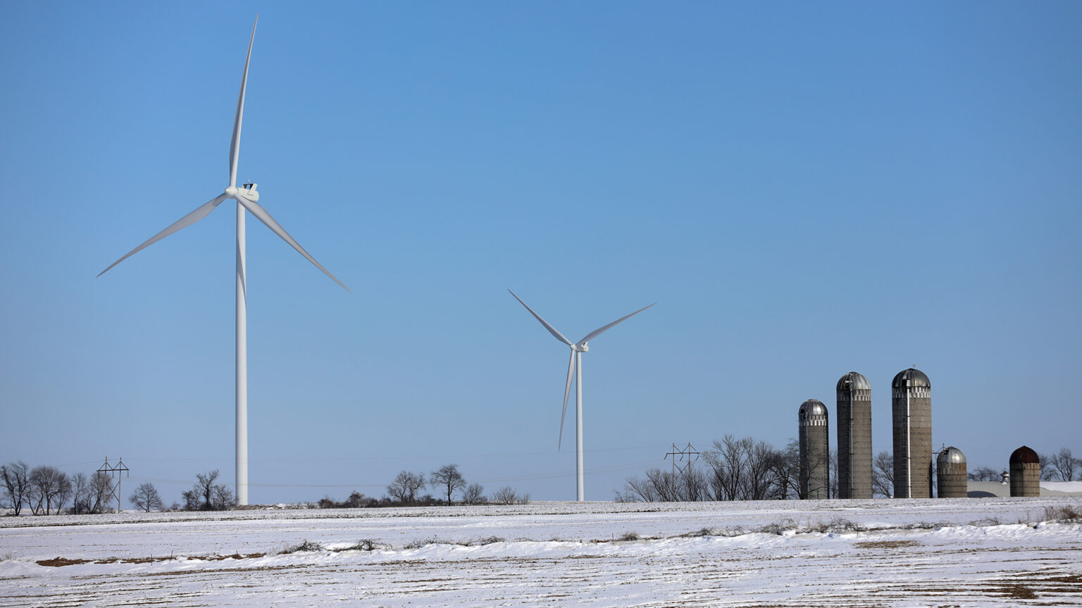 Two wind turbines stand in a snow-covered farm field, with multiple silos, leafless trees and a power line along the horizon. Two wind turbines stand in a snow-covered farm field, with multiple silos, leafless trees and a power line along the horizon.