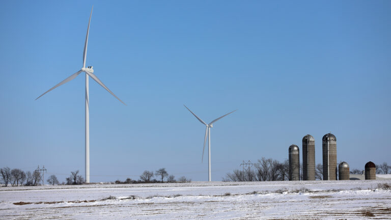 Two wind turbines stand in a snow-covered farm field, with multiple silos, leafless trees and a power line along the horizon.