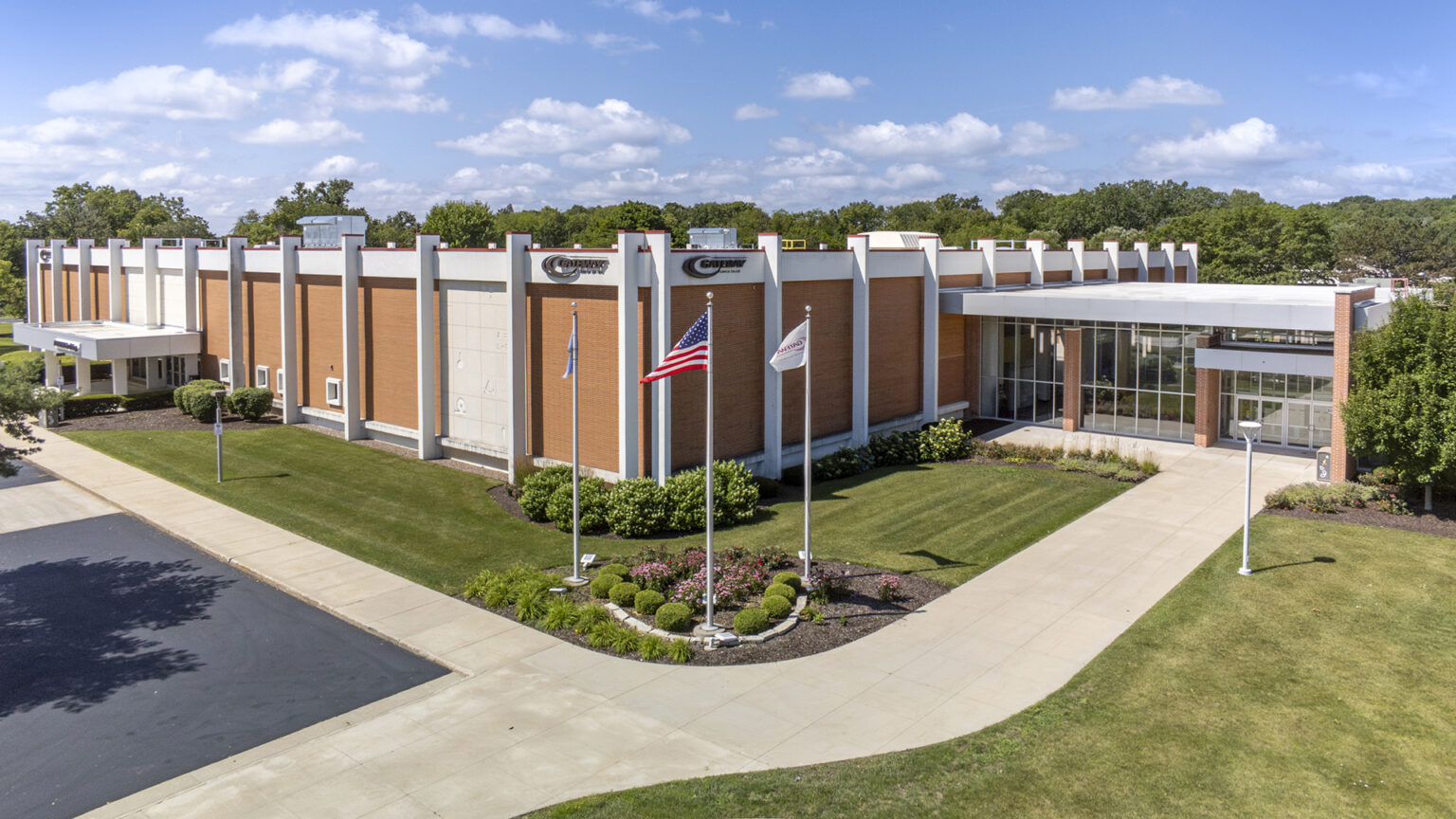 The U.S. flag and two other flags fly on flagpoles in a garden at the corner of two sidewalks in front of a brick building with multiple entrances, with lawns and a parking lot in the foreground and a line of trees on the horizon in the background under a partly cloudy sky.