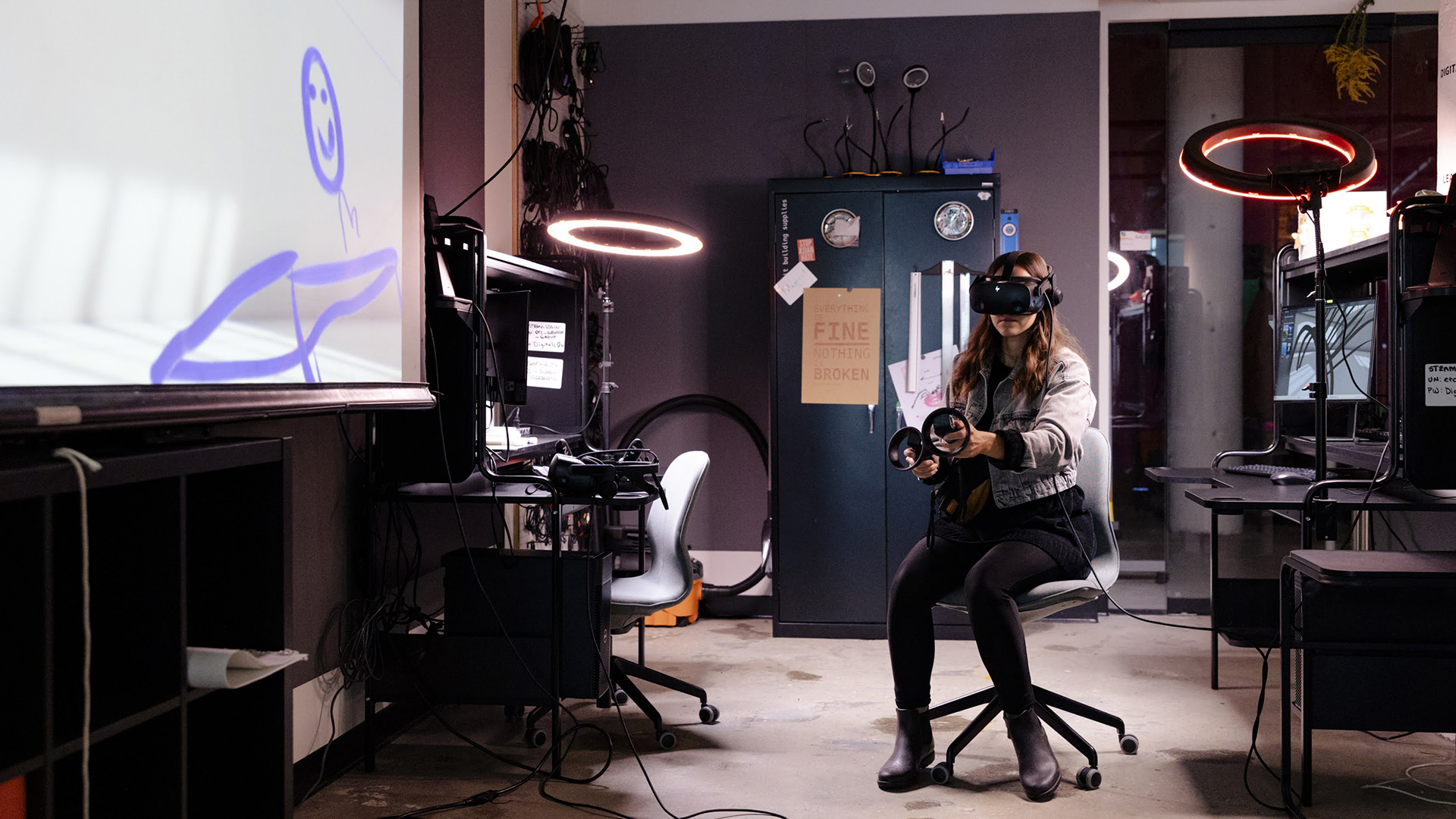 A student sits in a rolling chair while wearing virtual reality goggles and holding wired controllers in each hand in a room with desks, a metal cabinet, ring lights and a projector screen with a digital drawing displayed on its surface.