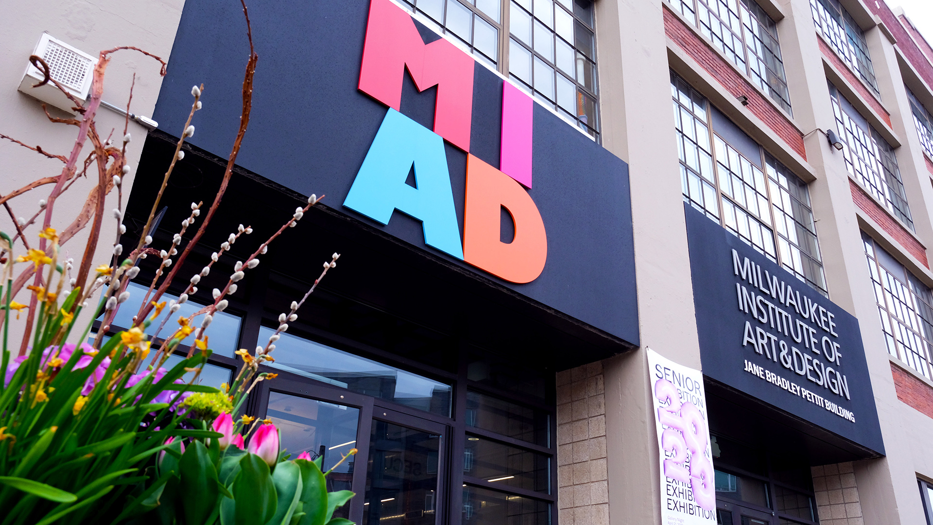 Letter signs reading "MIAD," "Milwaukee Institute of Art & Design," and "Jane Bradley Pettit Building" are affixed to the front of a multi-story building with a glass door entrance and multiple windows with small panes, with flower garden in the foreground.