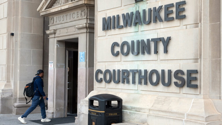 A man with a backpack walks onto a commercial floormat leading to the open wood doors of entrance outlined with pillars, a pediment and a bas relief sign reading JUSTICE in a masonry building with a letter sign reading Milwaukee County Courthouse on an adjacent wall and an enclosed trash receptacle in front of it.