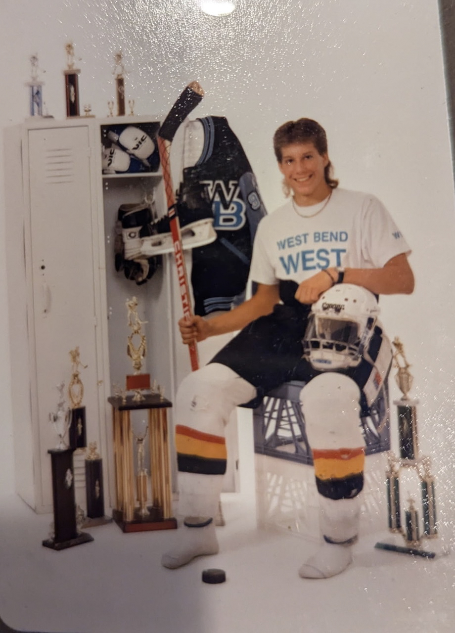 A light source reflects off the top of a matte-printed photograph showing a portrait of Scott Meyer wearing hockey pants and shin guards while seated with his left hand resting on a hockey helmet on his left leg and holding a hockey stick in his right hand, next to two lockers with a letter jacket hanging and hockey skates hanging on an open door and with multiple trophies and a hockey puck on the floor.
