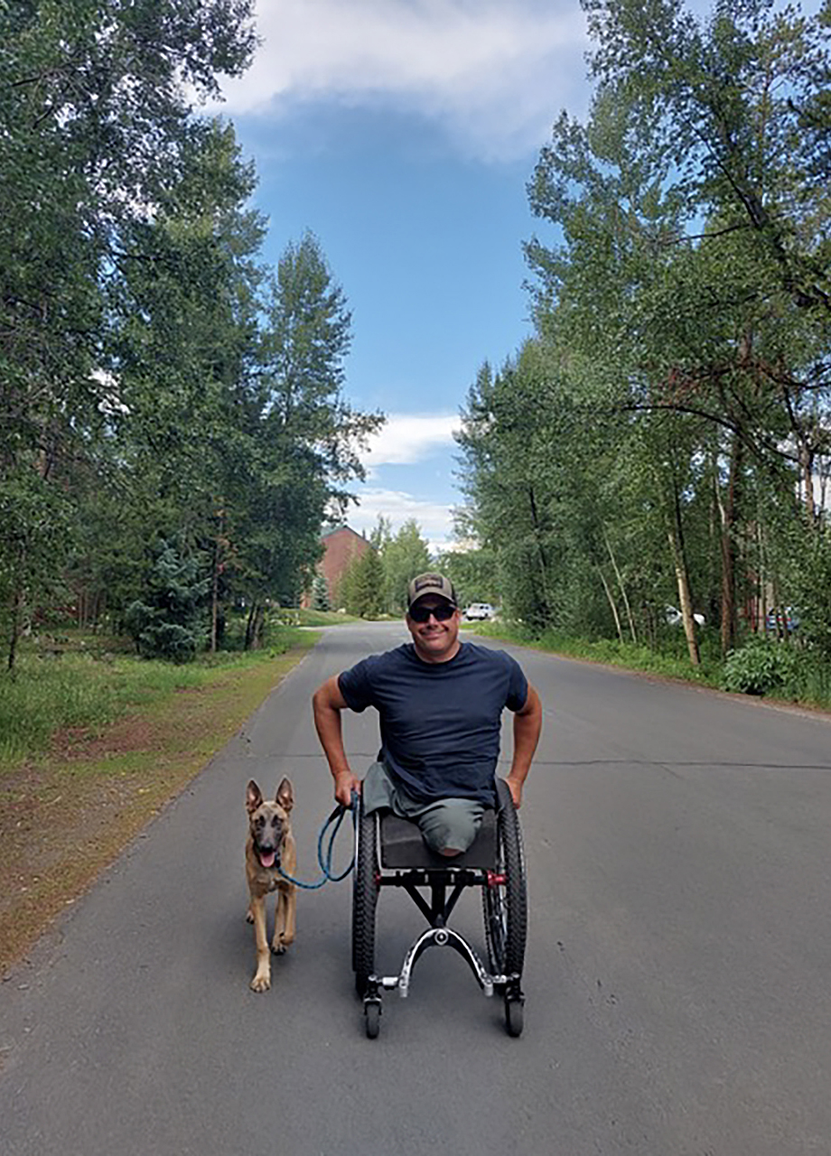 Scott Meyer poses for a photo and holds a leash in his right hand with a dog to his side while seated in a wheelchair on an asphalt road, with trees on either side and building in the background.