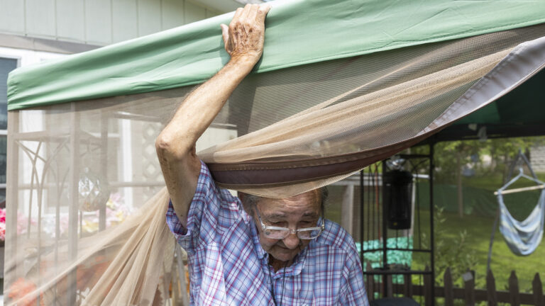 Jimmy Novy holds the top of a yard tent while sanding under a screen that has been drawn back to one side, with a building, fence, lawn and trees in the background.