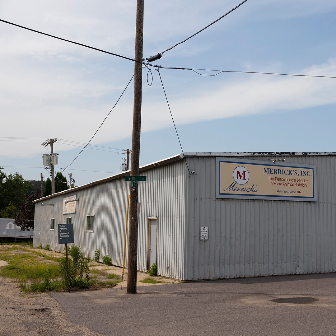 A utility pole connected to multiple wires stands next to the corner of a single story building with corrugated metal siding and a sign with the words "Merrick's, Inc." and "The Performance Leader in Baby Animal Nutrition" that is adjacent to a sidewalk overgrown with grass and with trees and other utility poles in the background.