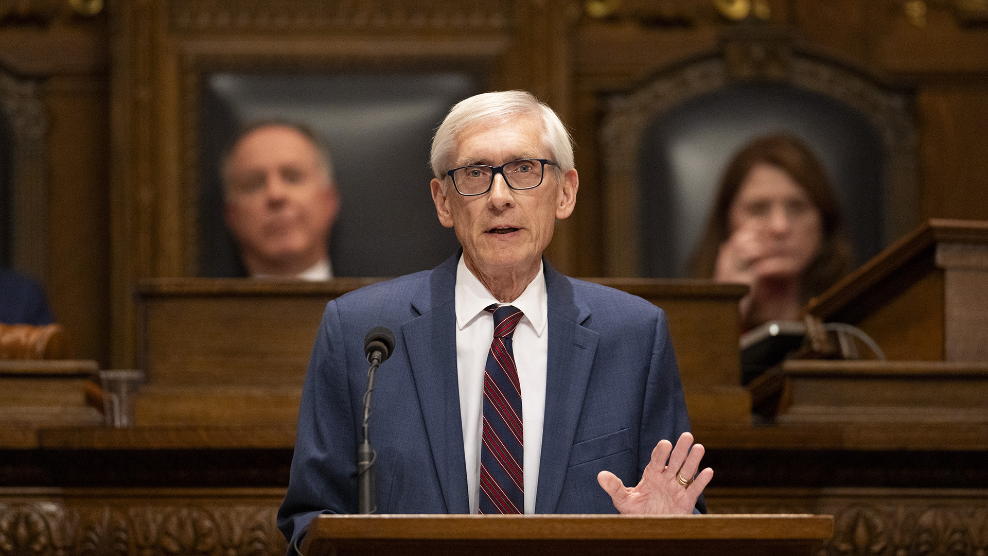 Tony Evers gestures with his left hand while standing and speaking into a microphone mounted to the top of a wood podium, with two out-of-focus people seated in high-backed leather and wood chairs on a wood legislative dais in the background.