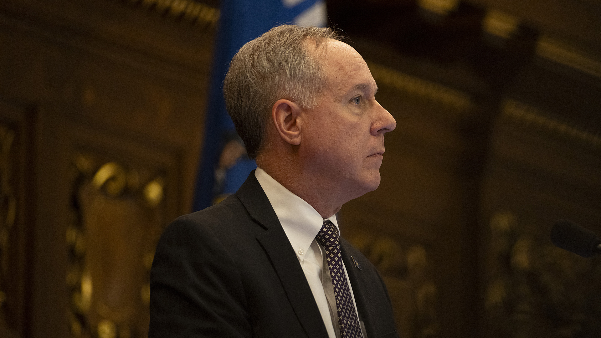 Robin Vos is seen in profile while standing with an out-of-focus Wisconsin flag and wood-paneled walls in the background.