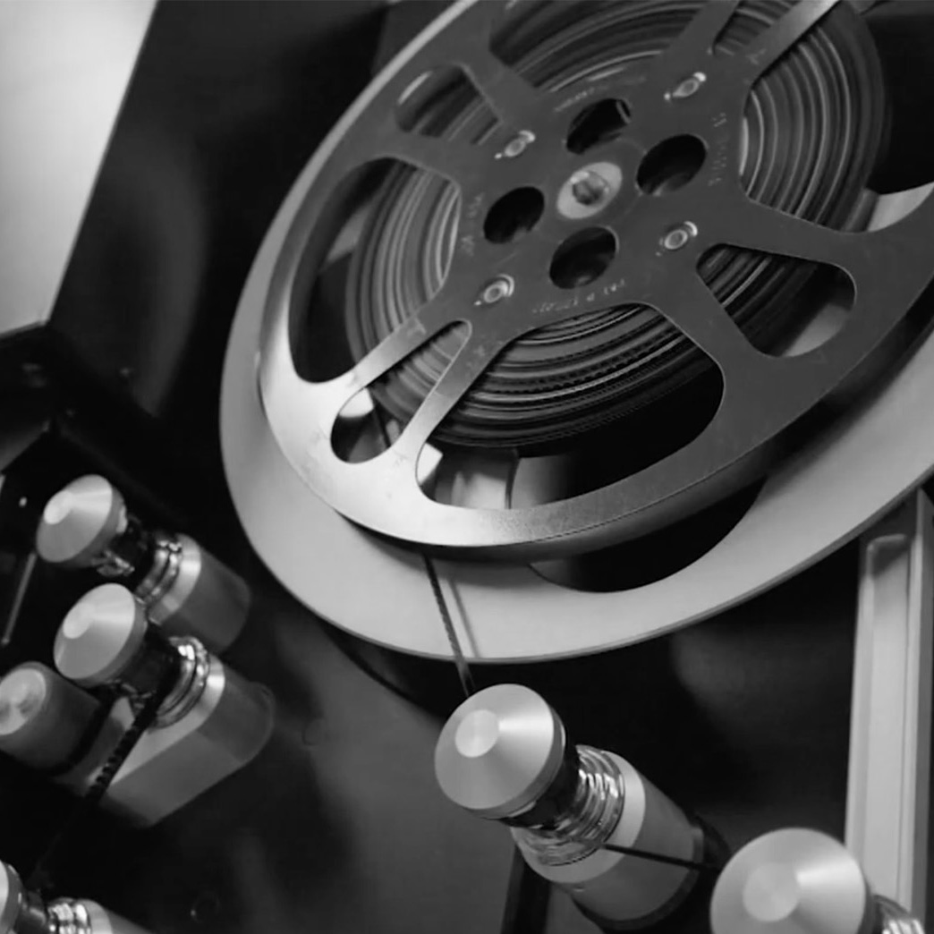 Close-up black-and-white photo of a reel-to-reel tape machine, showing a large metal reel wound with magnetic tape and a series of rollers and tension arms guiding the tape through the playback path.