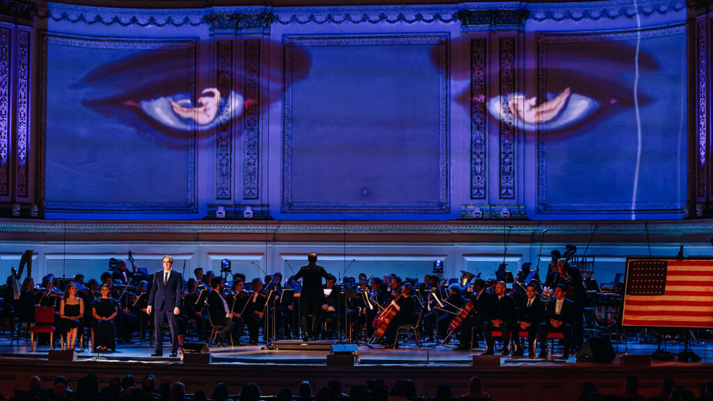 An orchestra performs on a darkened stage while a pair of eyes are projected onto the rear wall.