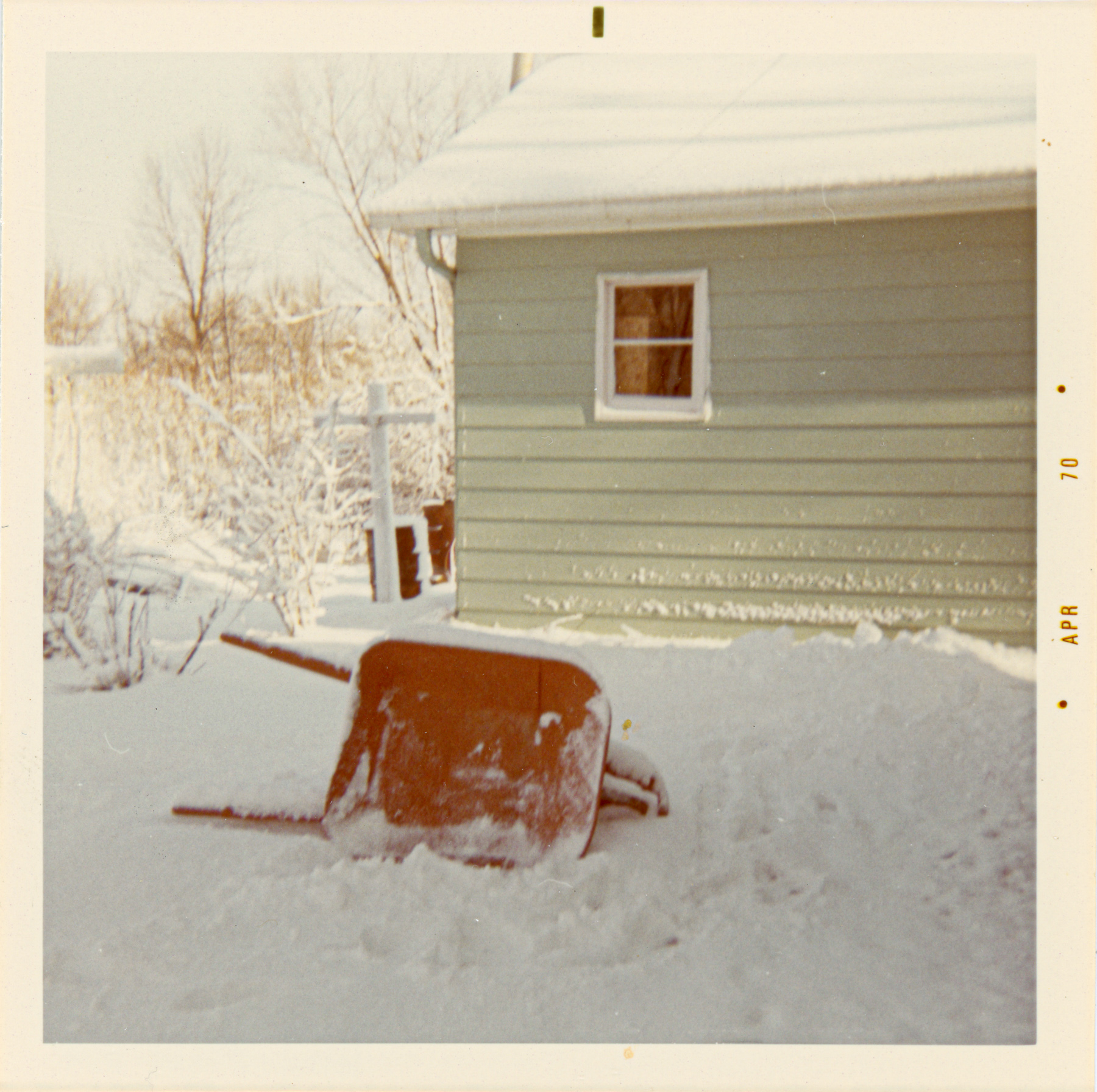 A red wheelbarrow tipped on its side in deep snow beside a pale green house. Bare trees and snow-covered branches fill the background. The photo border is marked “APR 70.”