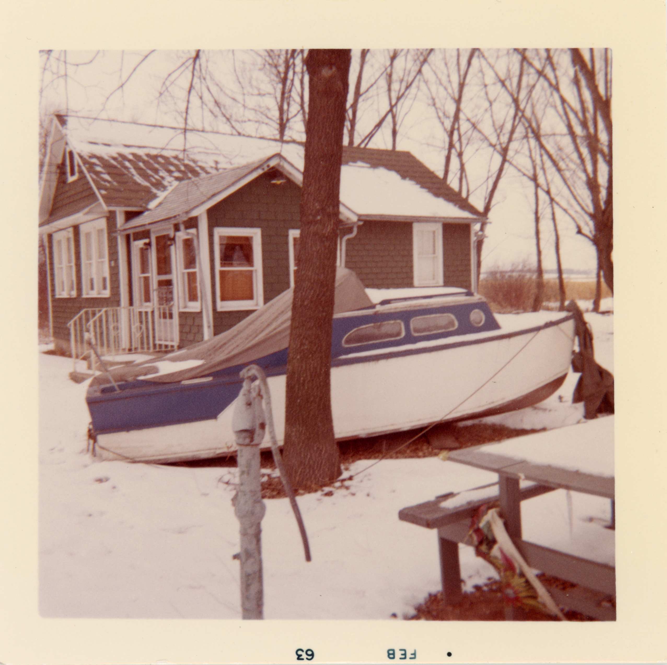 A small blue and white boat covered with a tarp sits on land beside a dark green house in winter. Snow covers the ground, and bare trees surround the house. A hand water pump and picnic table are visible in the foreground. The photo border reads “FEB 63."
