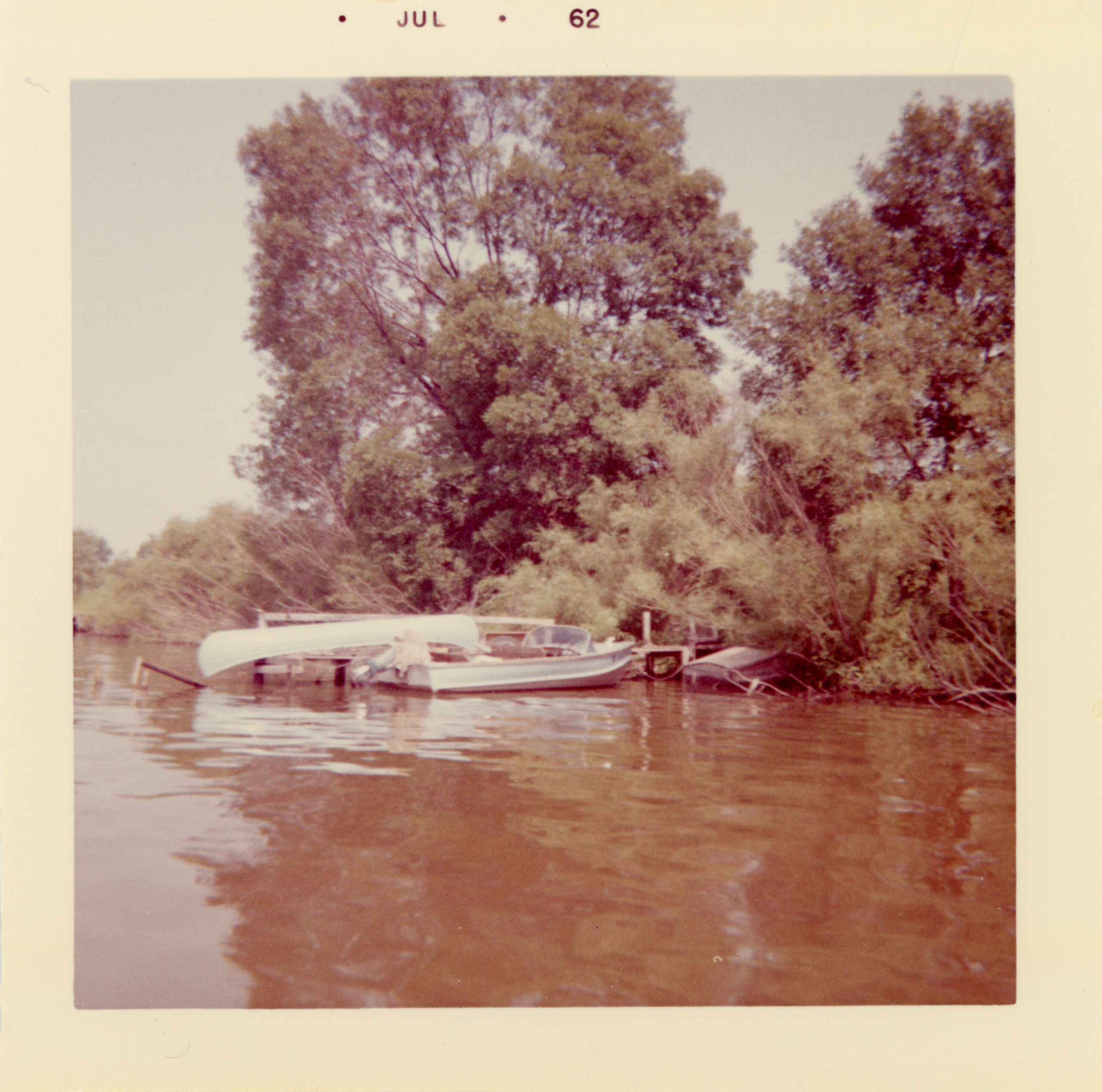 A small motorboat and a white canoe are tied to a wooden dock along a tree-lined riverbank. The water reflects the brown and green tones of the scene. The photograph has a warm tint and a white border labeled “JUL 62.”