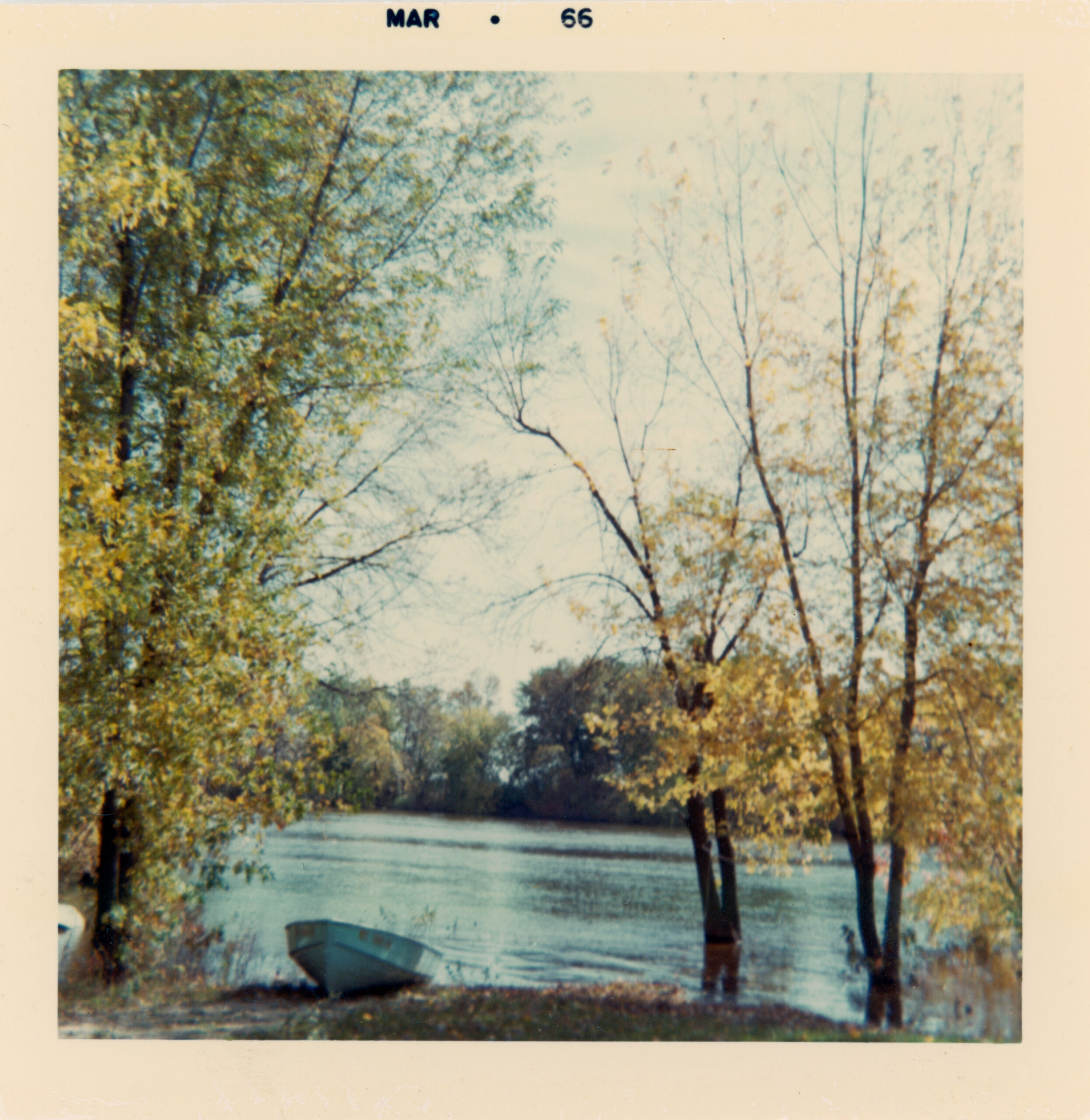 Color photograph dated March 1966 showing a calm river bordered by trees with sparse autumn leaves. A small rowboat rests on the grassy riverbank in the foreground, and sunlight filters through the branches over the still water.