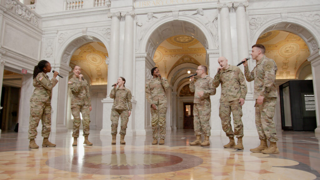 A chorus of soldiers wearing army fatigues stands in a semi-circle while holding microphones and singing.