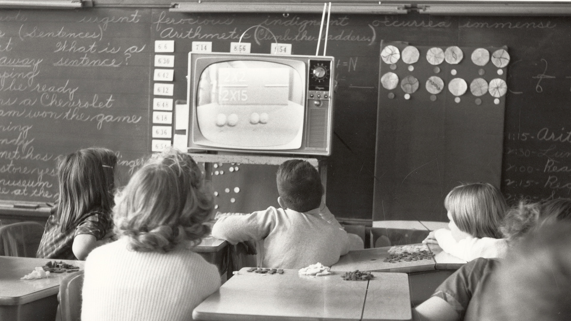 Elementary school students watch an educational television program in a classroom, likely from the 1950s or 1960s. A small black-and-white TV atop a cart displays math problems, while children seated at desks look on. A chalkboard behind them shows cursive writing and arithmetic lessons.