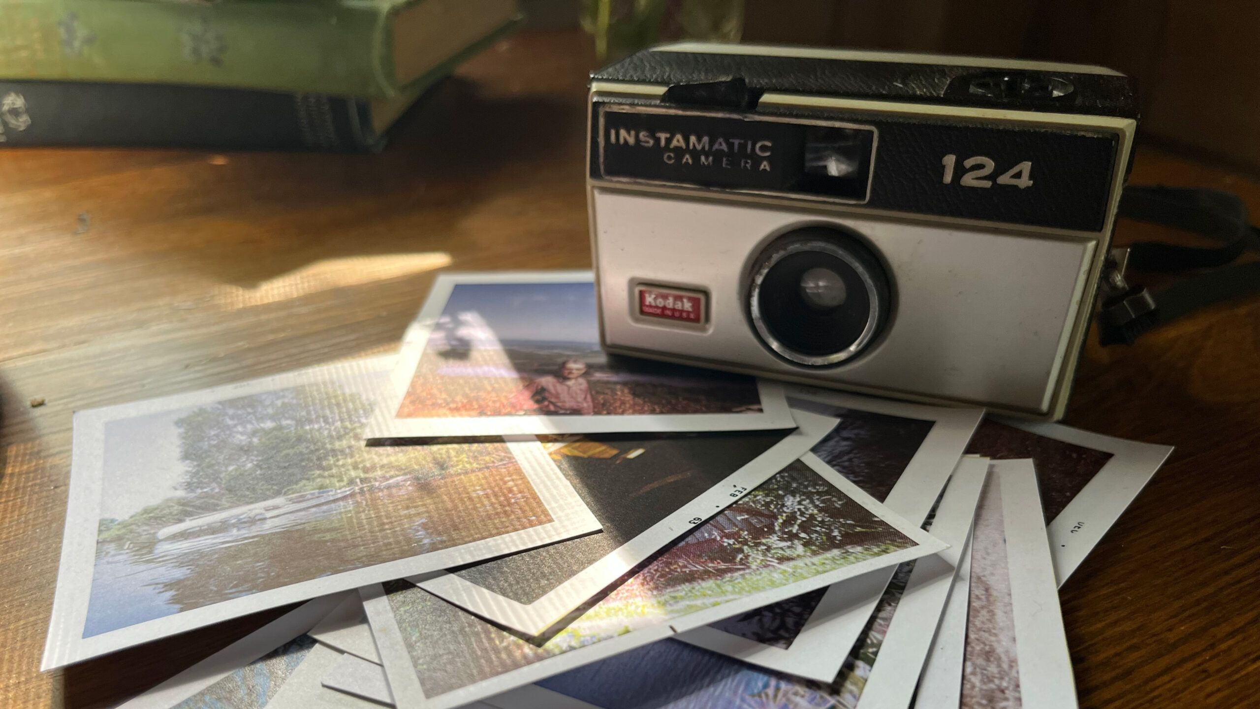 A vintage Kodak Instamatic 124 camera rests on a wooden table beside a small stack of printed color photographs. Sunlight filters across the table, casting a warm glow on the camera and photos, with a few old books blurred in the background.