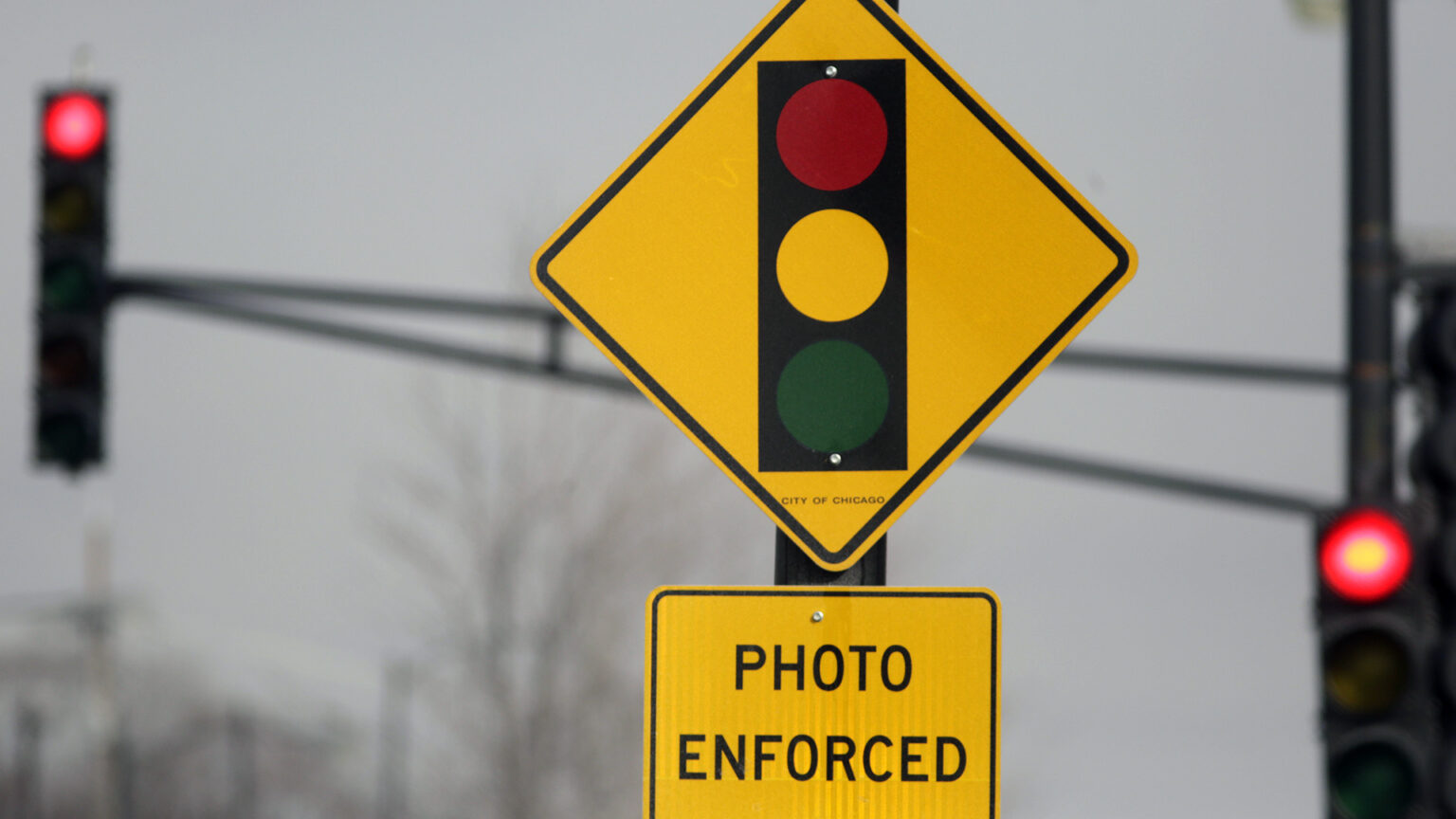 A diamond-shaped metal traffic sign with a graphic of a vertical traffic light and a rectangular metal traffic sign with the words Photo Enforced stand on a metal poll, with an out-of-focus traffic signal with two illuminated red lights in the background. A diamond-shaped metal traffic sign with a graphic of a vertical traffic light and a rectangular metal traffic sign with the words Photo Enforced stand on a metal poll, with an out-of-focus traffic signal with two illuminated red lights in the background.