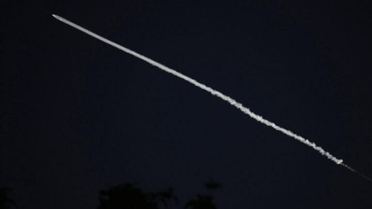 A rocket and its contrail arc from lower right to upper left across a dark nighttime sky.