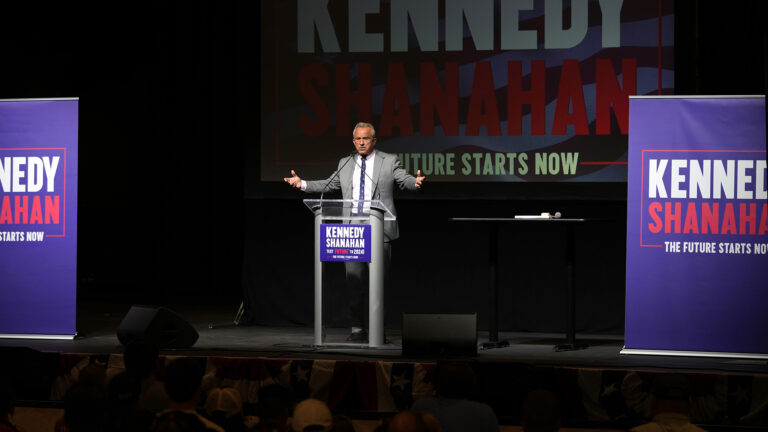 Robert F. Kennedy Jr. gestures with both arms held to the side while standing behind a plastic podium with two mounted microphones and a campaign sign on its front reading Kennedy Shanahan, with multiple larger signs to the side and in the background on a stage, with people seated below facing him. Robert F. Kennedy Jr. gestures with both arms held to the side while standing behind a plastic podium with two mounted microphones and a campaign sign on its front reading Kennedy Shanahan, with multiple larger signs to the side and in the background on a stage, with people seated below facing him.