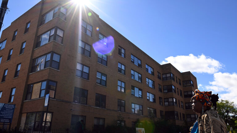 A pedestrian walks in front of a five-story building with brick walls and small glass windows, with the sun shining over the edge of its roof.