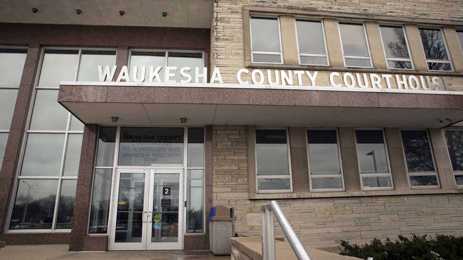 A letter sign reading Waukesha County Courthouse stands on the top of an awning with masonry panels that stands above a glass double-door entrance to a multistory building with glass and different types of masonry and multiple rows of windows.