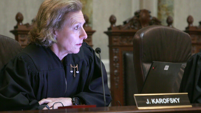 Jill Karofsky sits in a high-backed leather chair at a judicial bench with a nameplate reading J. Karofsky, with a row of empty high-backed wood and leather chairs in a room with marble pillars and masonry.