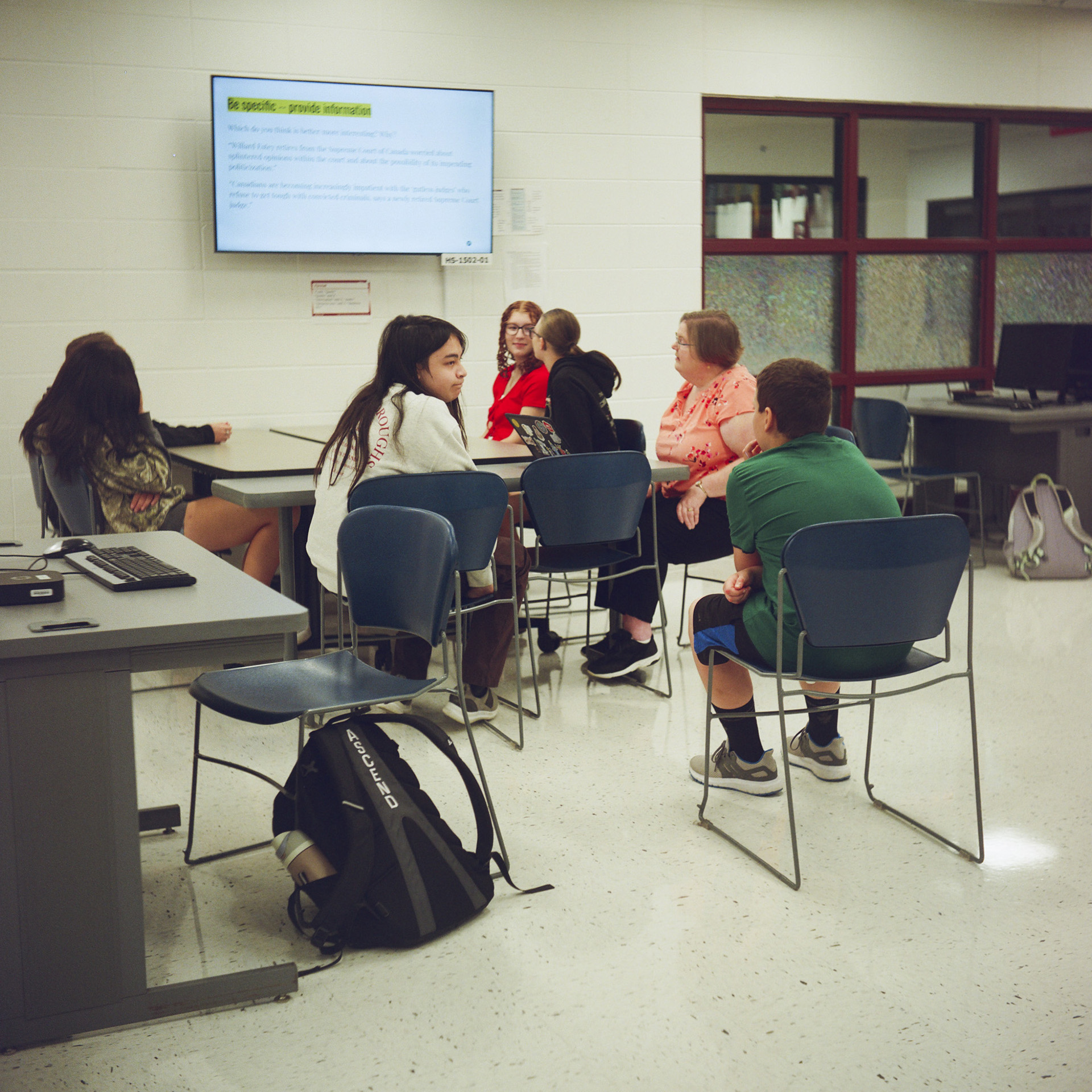 Six students and a teach sit in stackable plastic chairs around and near two tables and face a monitor mounted on a painted concrete block wall, in a room with a vinyl tile floor and a set of windows with transparent and glazed glass panels.