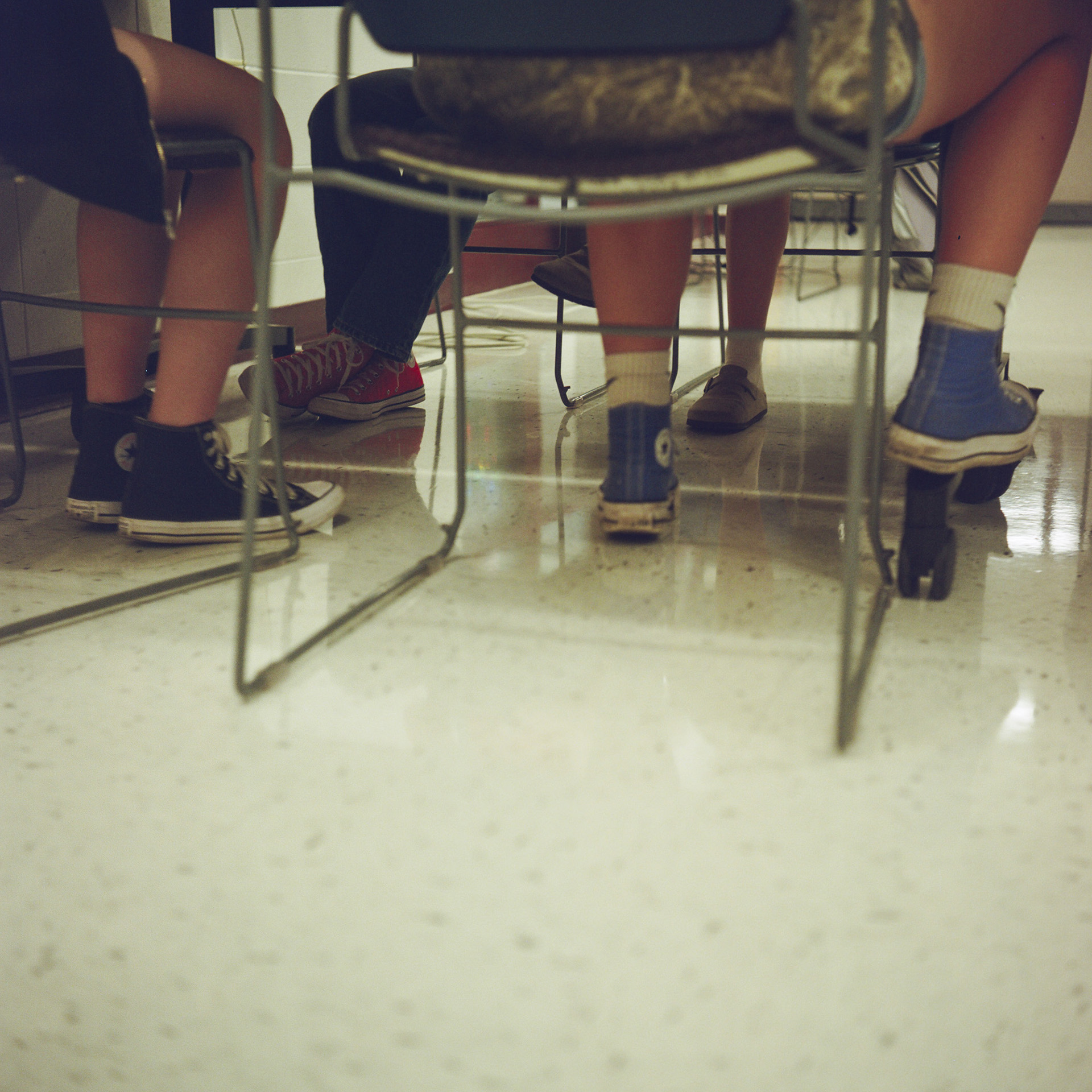 The feet of three students wearing Converse high top shoes rest of a vinyl panel floor and the metal legs of a table.