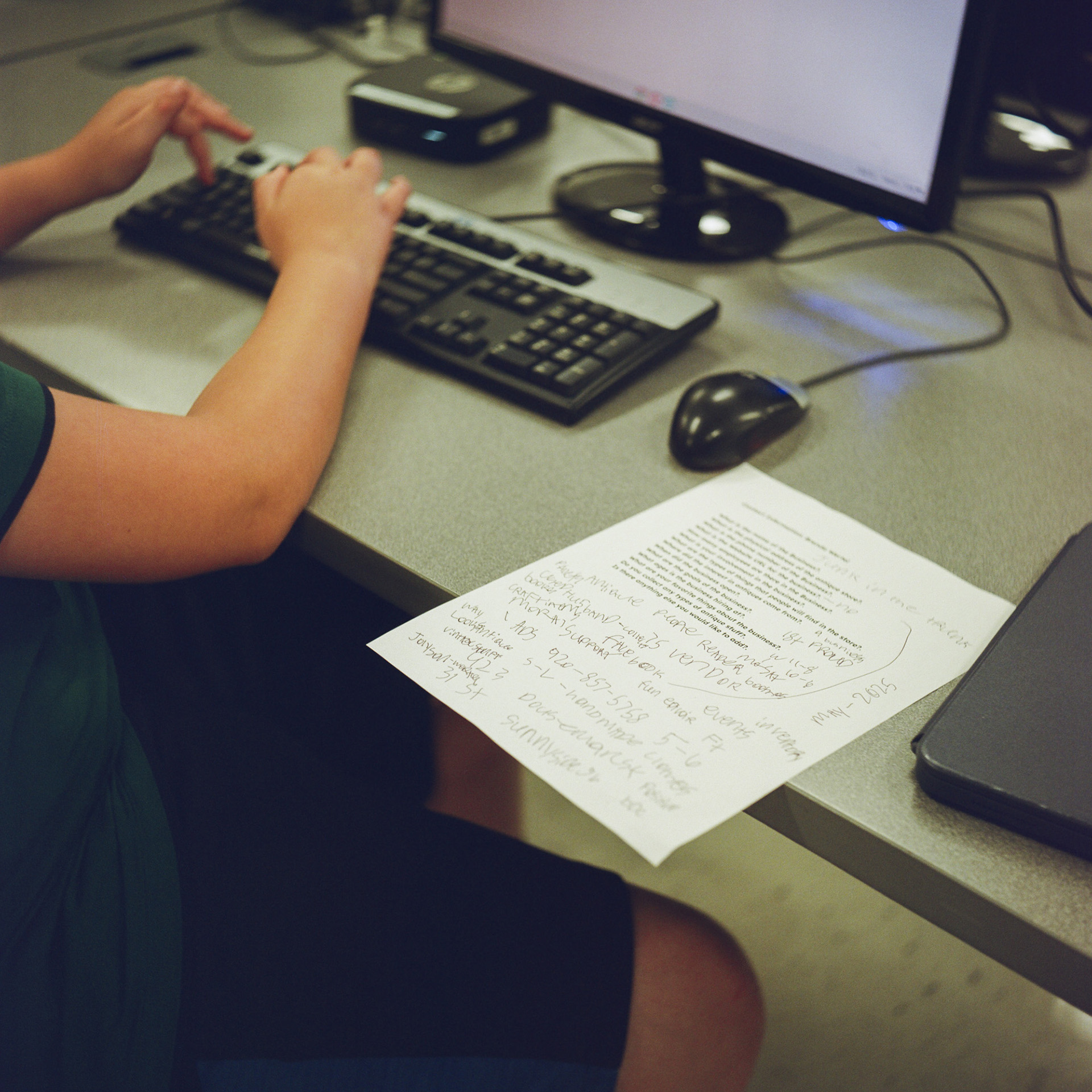 The hands of Daniel Roggenbauer type on a keyboard in front of a computer monitor and next to a wired mouse and a printed sheet of paper with handwritten notes on the top of a table.